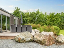 Una terraza moderna al aire libre con mobiliario de mimbre y un paraguas grande, rodeada por un paisaje natural con rocas y follaje.