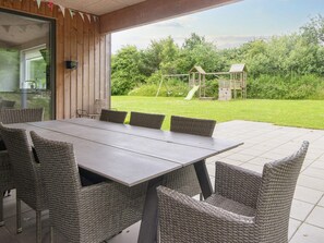 Terrassa exterior amb taula de dinar i cadires de vimet, amb vistes a una zona enjardinada i una zona de jocs per a infants.
