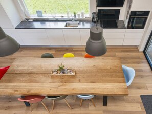 A wooden dining table with a tray of decorative items, surrounded by colorful chairs, in a modern kitchen with white cabinets and a window view.