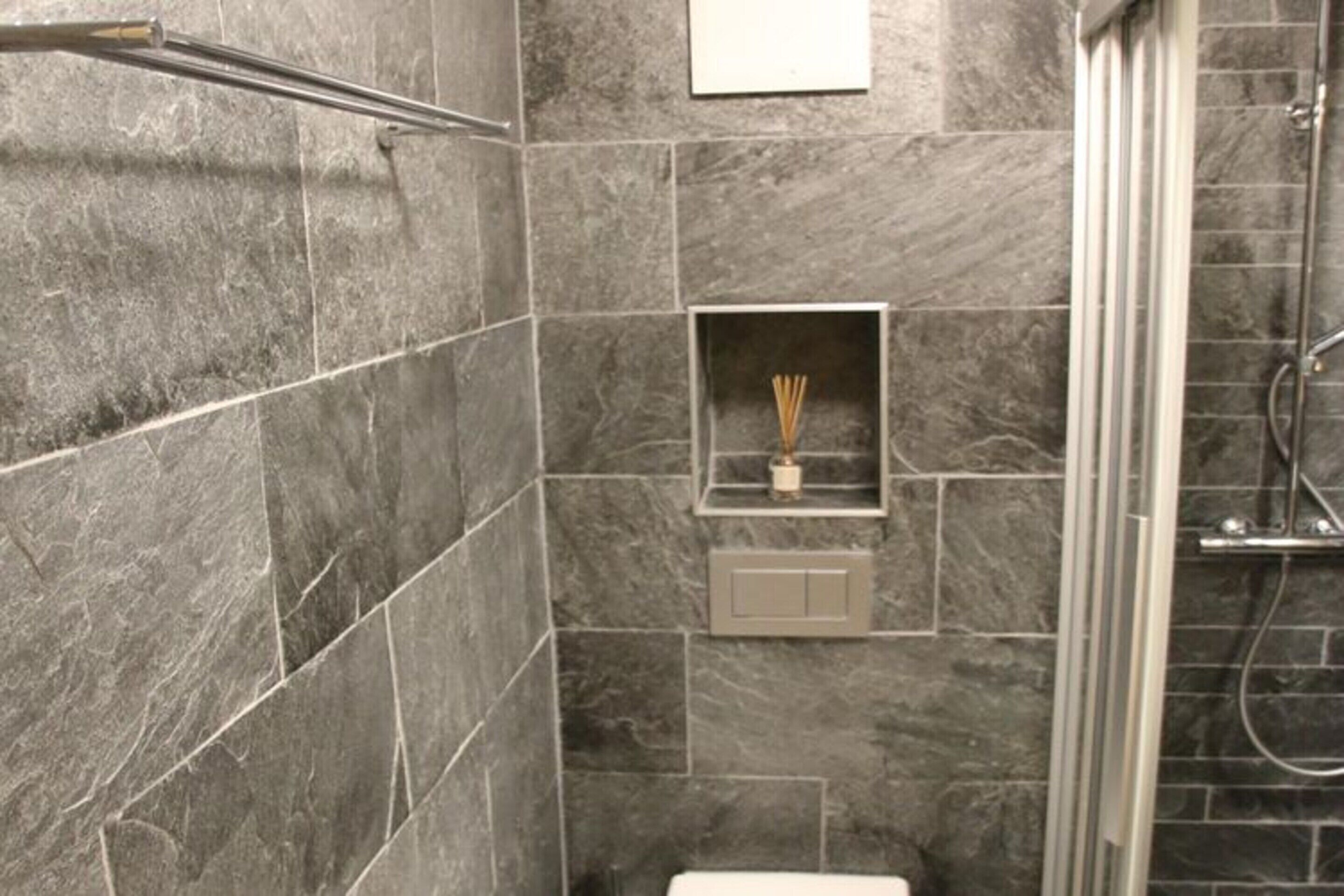 A bathroom with grey marble tiles, a shower area, and a small shelf with a bottle and reed diffuser.