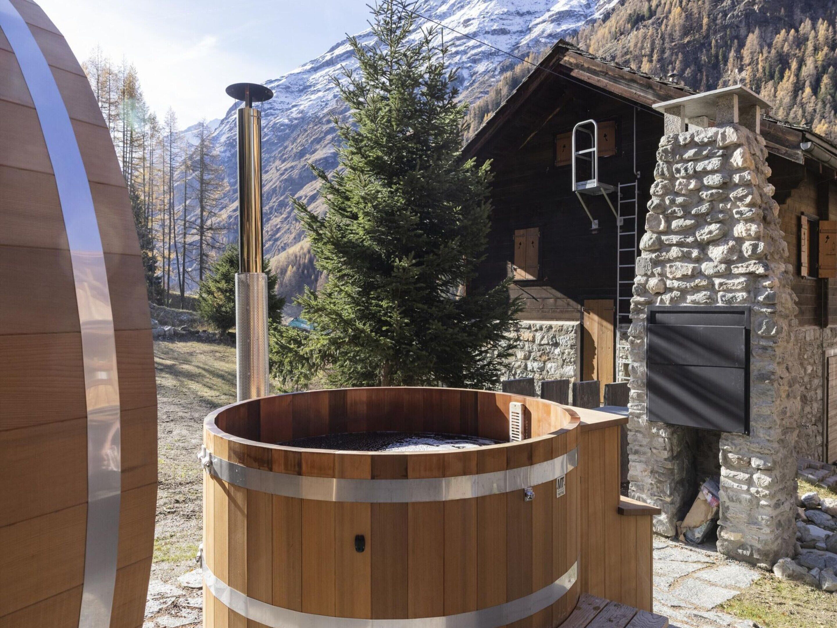 Un jacuzzi de madera en una zona montañosa, con un edificio de piedra y una cabaña de madera al fondo.
