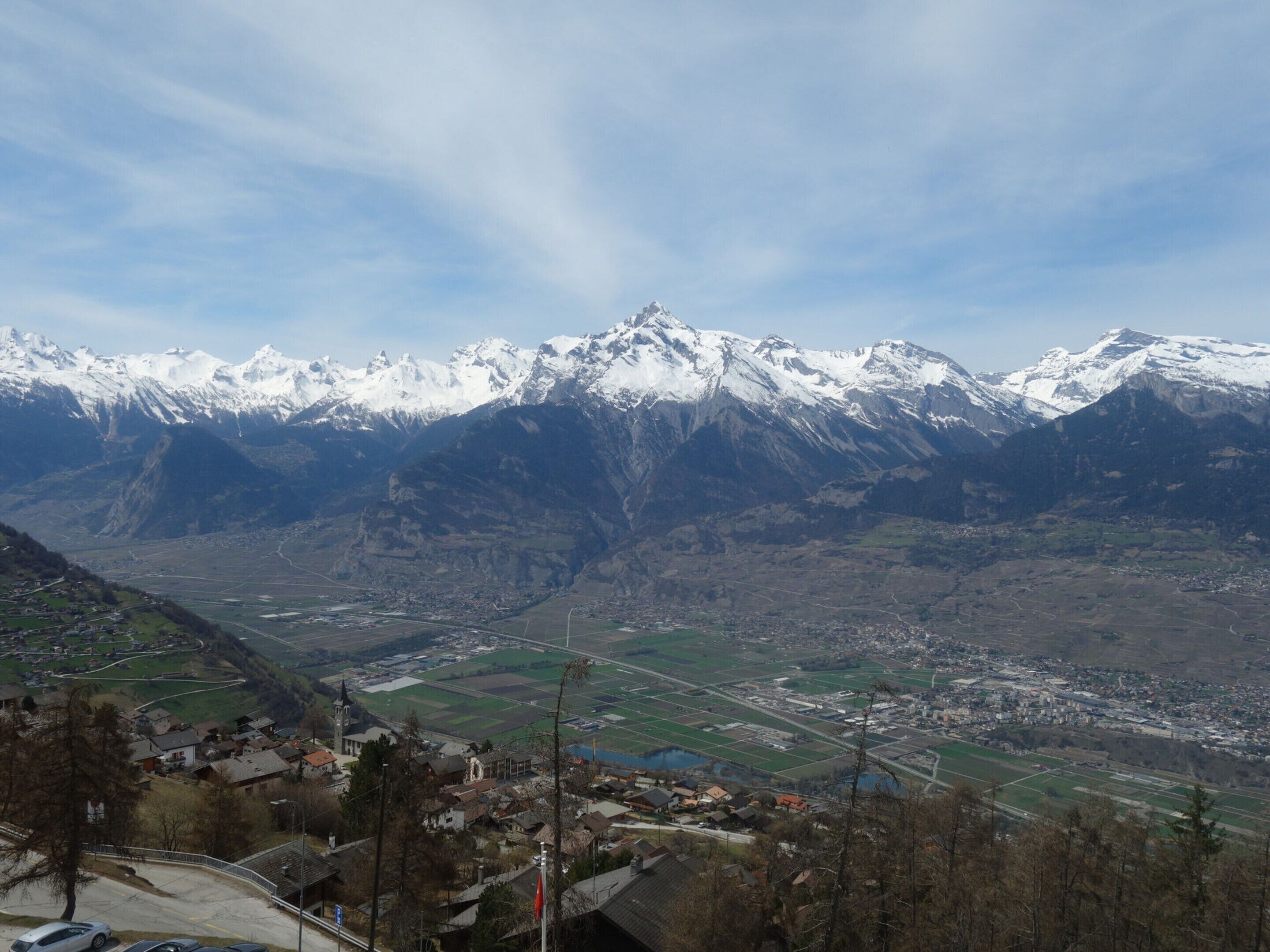 A mountainous landscape with snow-capped peaks, a valley with fields, and a small town with buildings and roads.