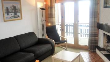 A living room with a black sofa, a wooden chair, a small white table, a fireplace, and a balcony with a railing.