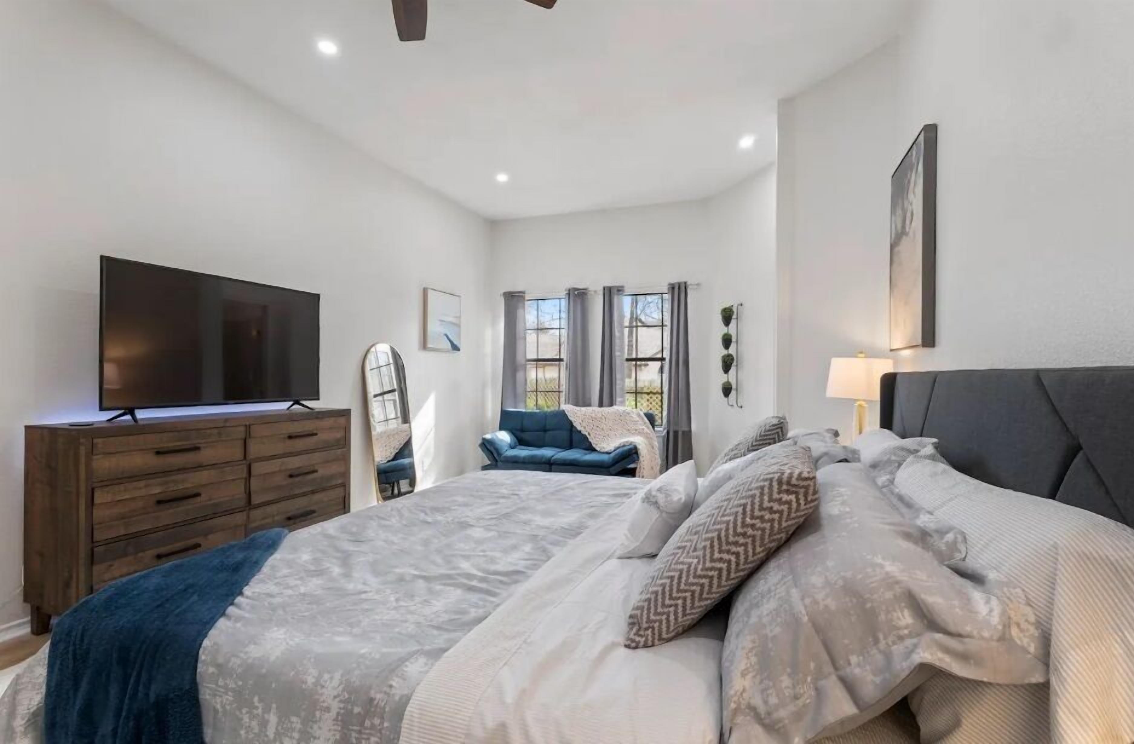 Un dormitorio con una cama grande, un televisor sobre una cómoda de madera, un sofá azul y una ventana con cortinas.