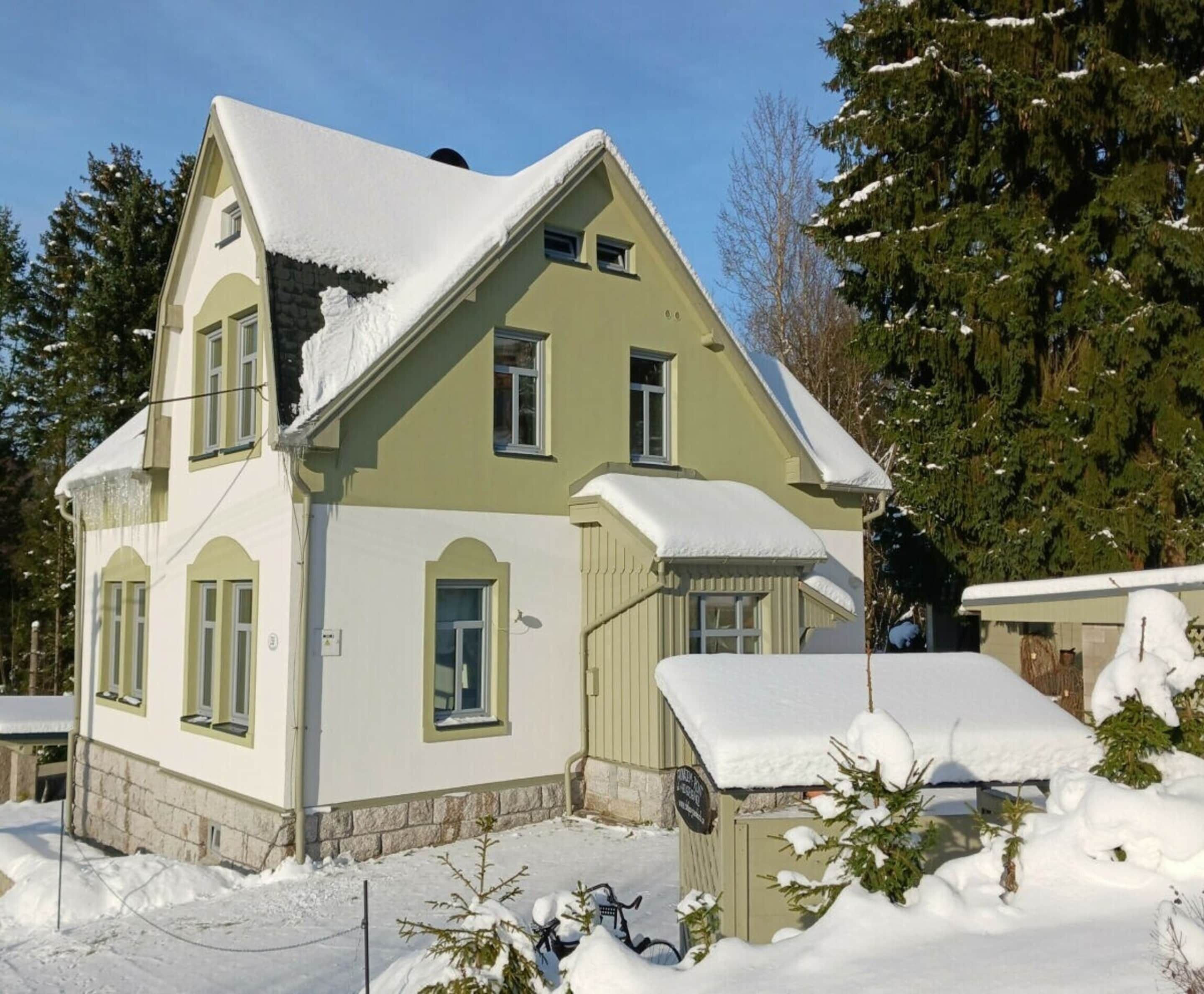 A two-story house with a steep roof, surrounded by snow-covered trees.