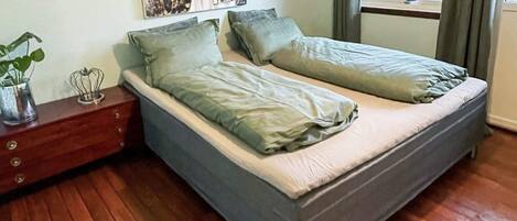 A bedroom with a double bed, a wooden nightstand, a small table with a plant, and a window with curtains.