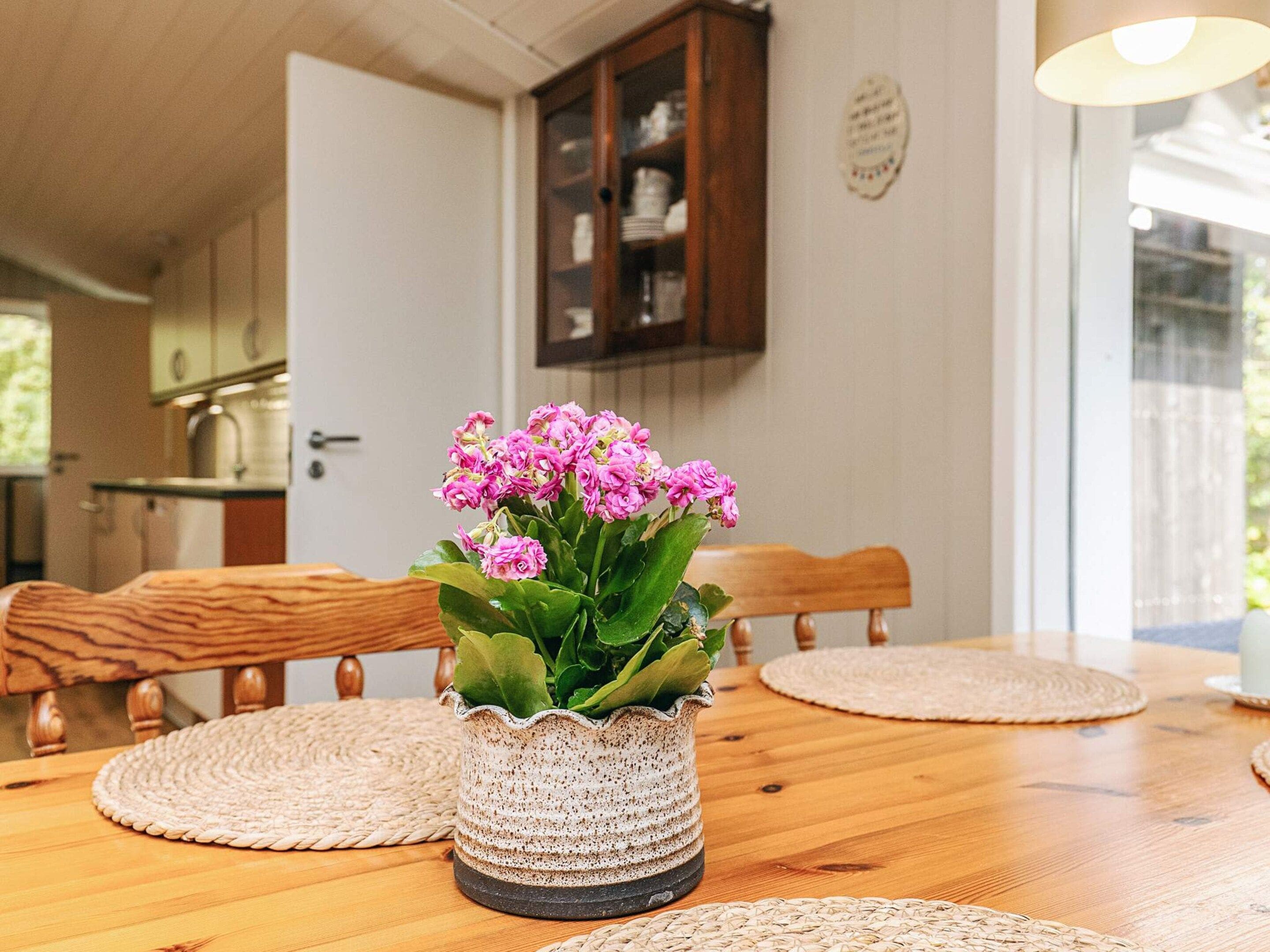 Una mesa de comedor con un mantel tejido, una maceta de cerámica con flores rosas, sillas de madera y una cocina al fondo.