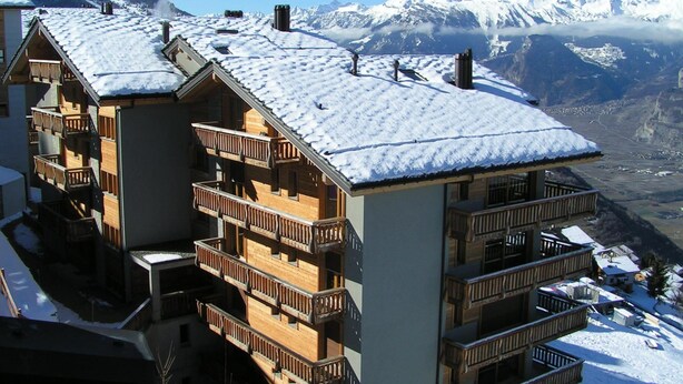Ein mehrstöckiges Chalet mit Holzbalkonen und einem mit Schnee bedeckten Dach, vor einer Kulisse aus schneebedeckten Bergen.