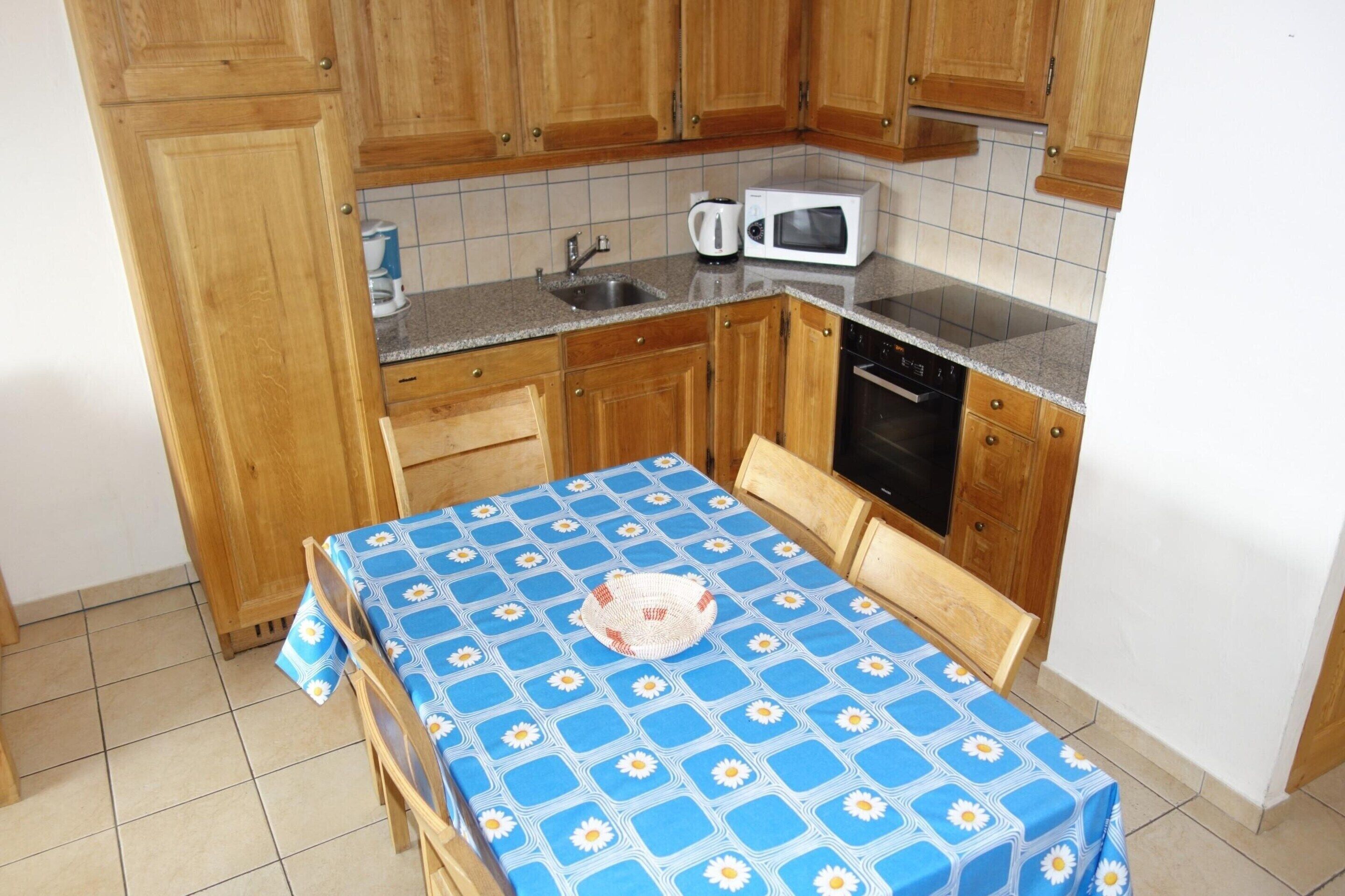 A small kitchen with wooden cabinets, a microwave, a coffee maker, and a table with a blue tablecloth.