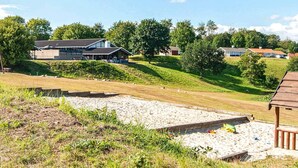 A residential area with a modern house, a sandy area with a wooden structure, and a grassy hill with trees. - 5 Person Holiday Home in Aabenraa (Aabenraa)