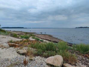 Un paysage côtier caractérisé par une rive rocheuse, une végétation clairsemée et une vue lointaine d’un voilier sur l’eau.