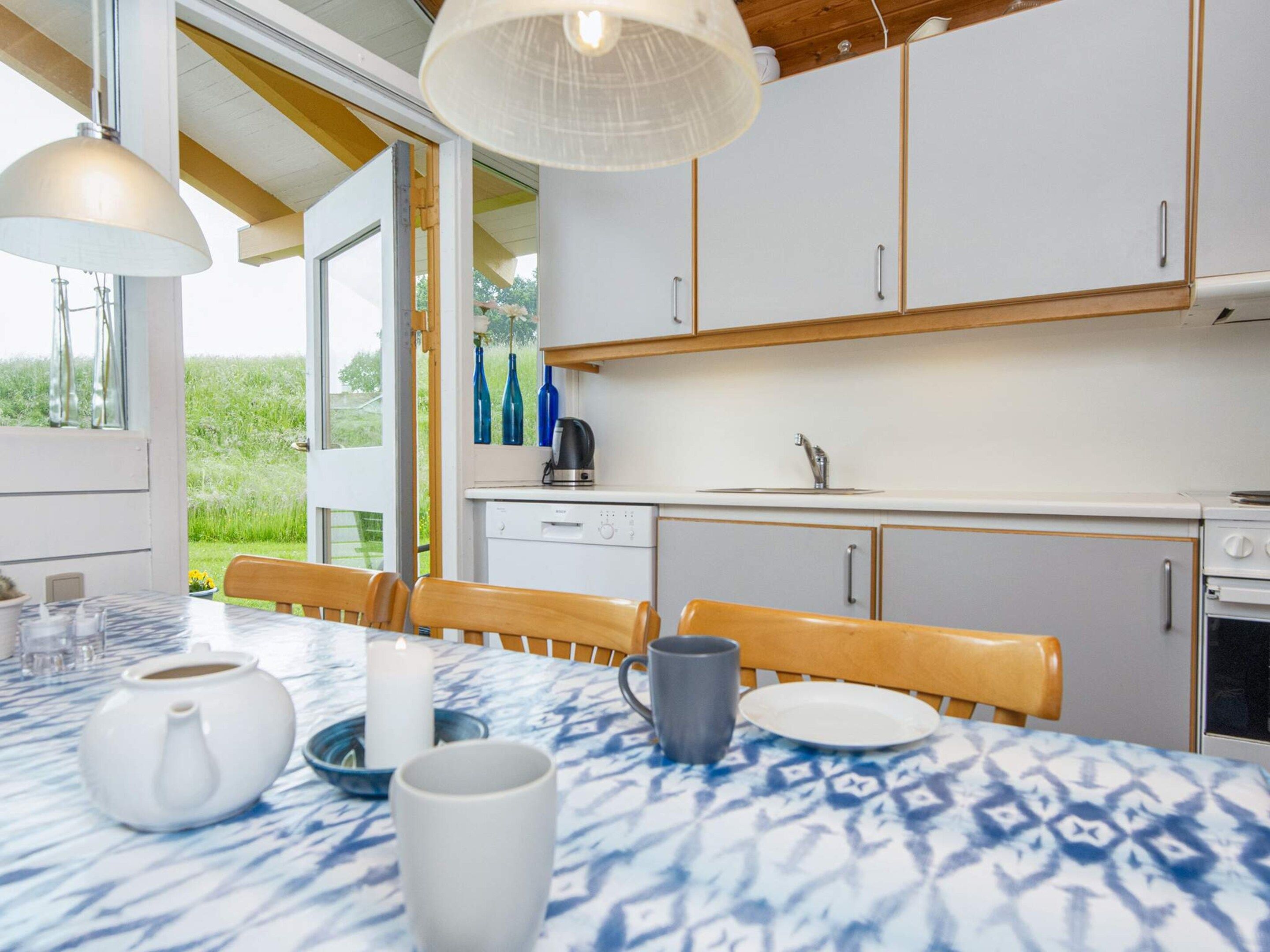 A modern kitchen with white cabinets, a dining table with a blue patterned tablecloth, and a view of greenery outside.