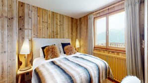 Ein schlafreicher Raum mit Holzwandverkleidung, einem Bett, zwei Nachttischlampen, einem Fenster mit Vorhängen und Blick ins Freie.