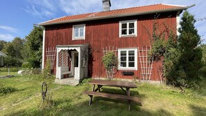 A red wooden house with a white door and windows, a wooden bench, and a garden area.