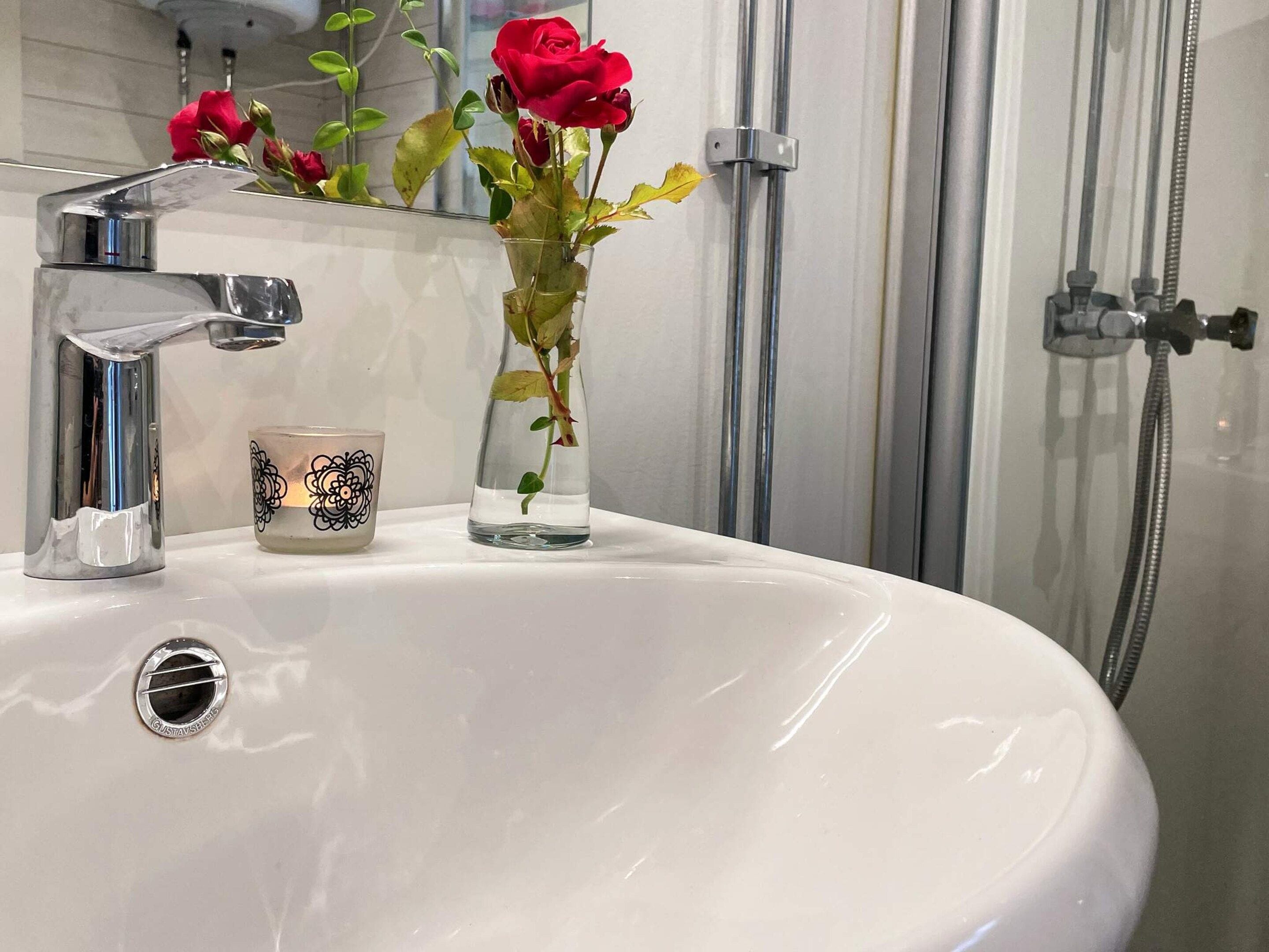 A bathroom with a white sink, a chrome faucet, a glass vase with a red rose, and a candle with a black floral design.