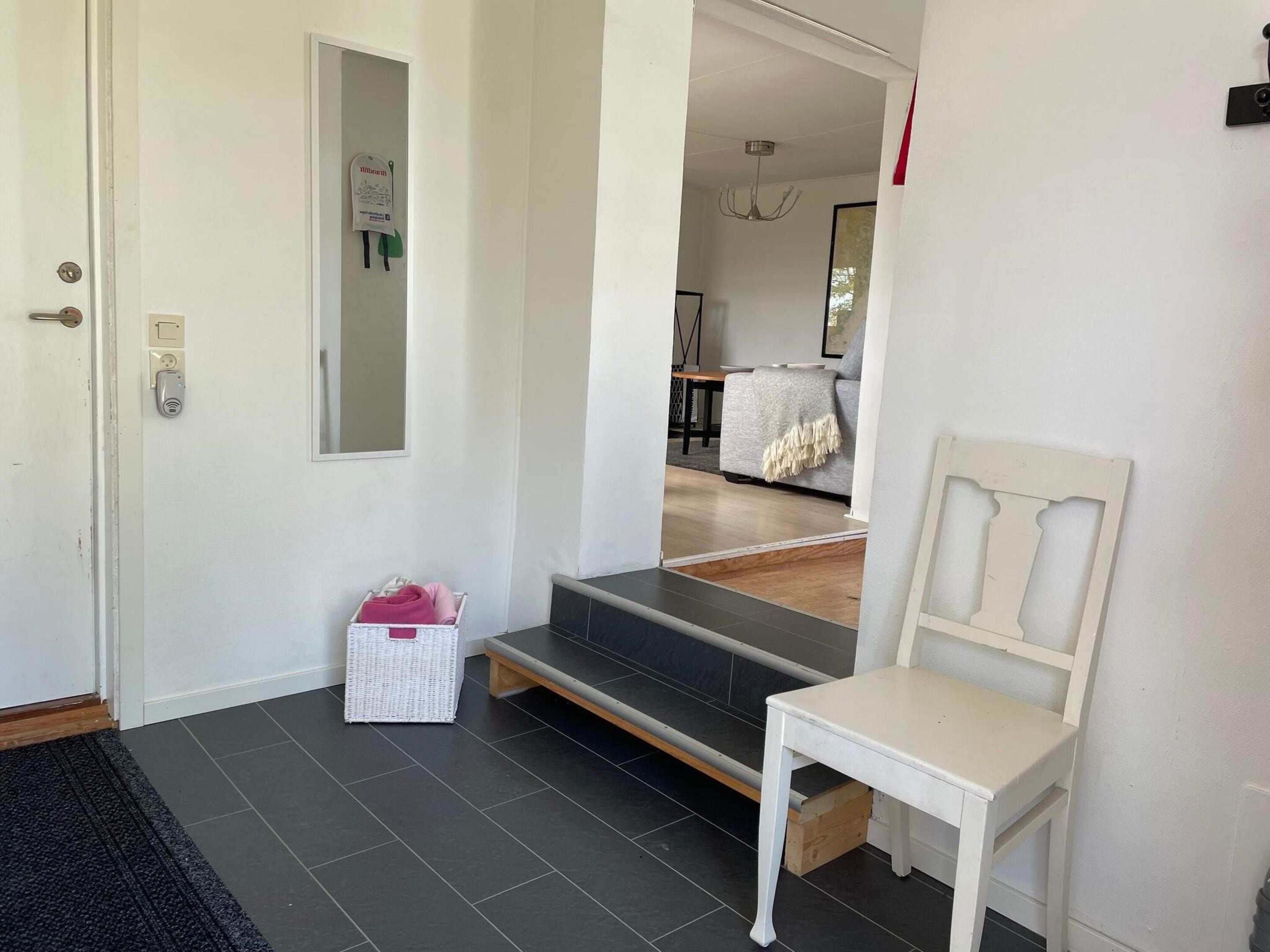 A clean, modern entryway with a white door, a small step leading to a living area, and a white chair by the wall.