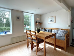 A room with a wooden dining table, chairs, a bench with cushions, a radiator, a window with a view of greenery, and a wall-mounted shelf with a radio. - 4 Person Holiday Home in Figeholm (Figeholm)
