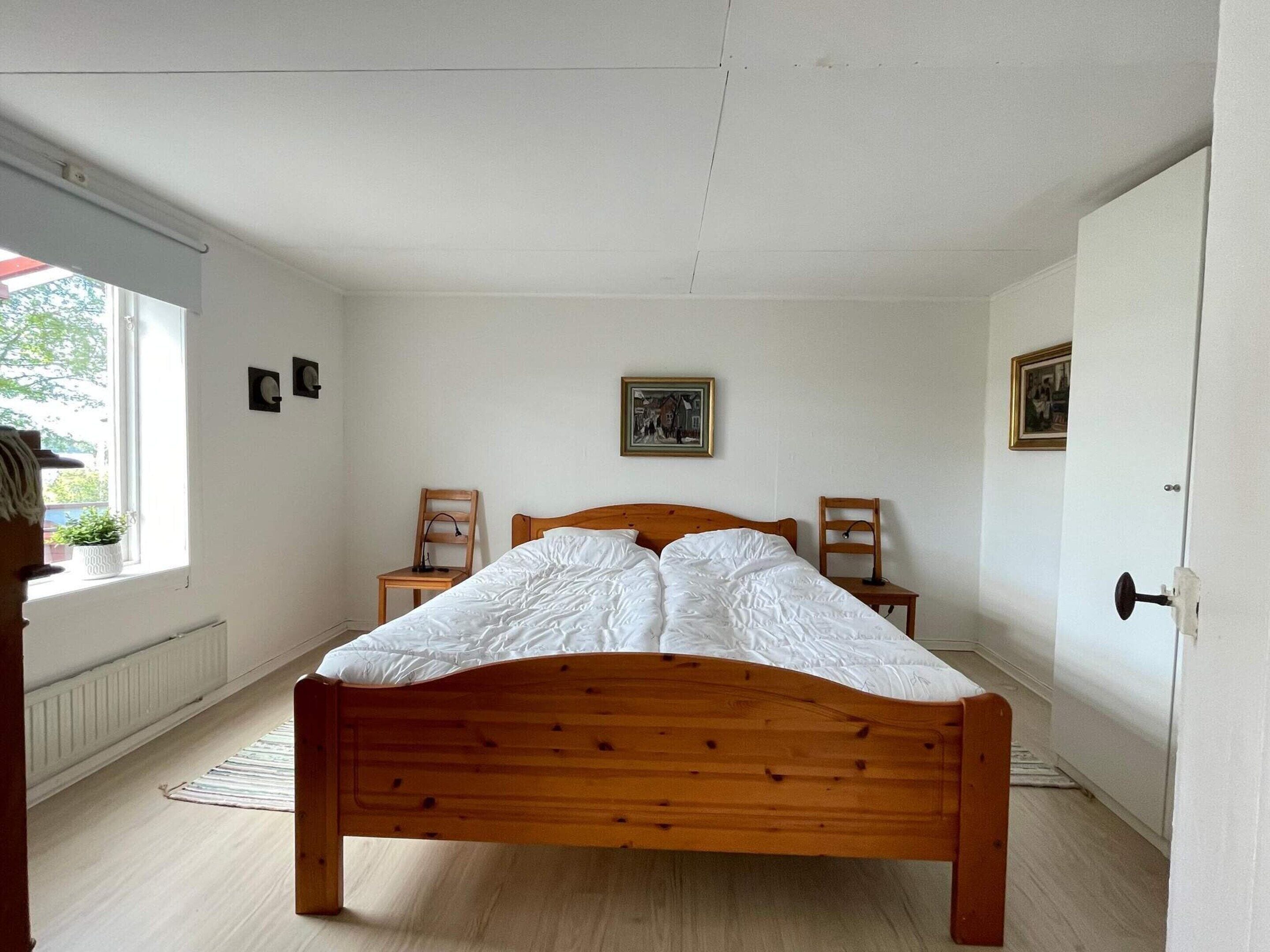 Ein Schlafzimmer mit einem Holzbett, zwei Stühlen, einem Heizkörper und einem Fenster mit Blick auf Grünflächen.
