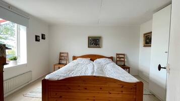 Ein Schlafzimmer mit einem Holzbett, zwei Stühlen, einem Heizkörper und einem Fenster mit Blick auf Grünflächen.