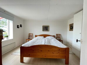 A bedroom with a wooden bed, two chairs, a radiator, and a window with a view of greenery.