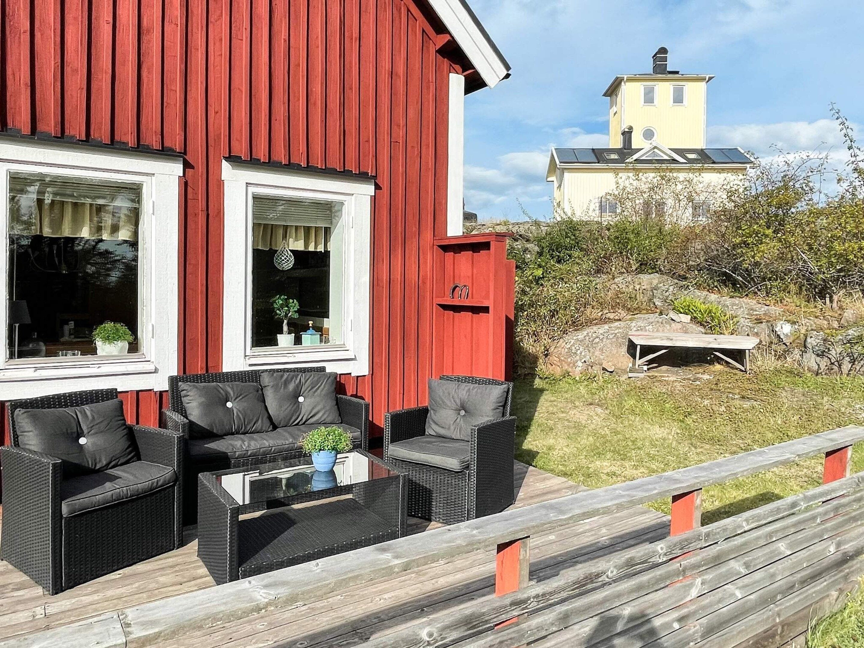 Ein rotes Gebäude mit zwei Fenstern, eine Holzterrasse mit Gartenmöbeln und ein Blick auf ein gelbes Gebäude im Hintergrund.