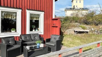Un edificio rojo con dos ventanas, una terraza de madera con mobiliario exterior y una vista de un edificio amarillo al fondo.