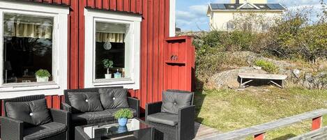 Ein rotes Gebäude mit zwei Fenstern, eine Holzterrasse mit Gartenmöbeln und ein Blick auf ein gelbes Gebäude im Hintergrund.
