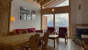 A dining area with a wooden table and chairs, a fireplace, and a view of mountains through large windows. - Pleasant Apartment With Balcony (Veysonnaz)