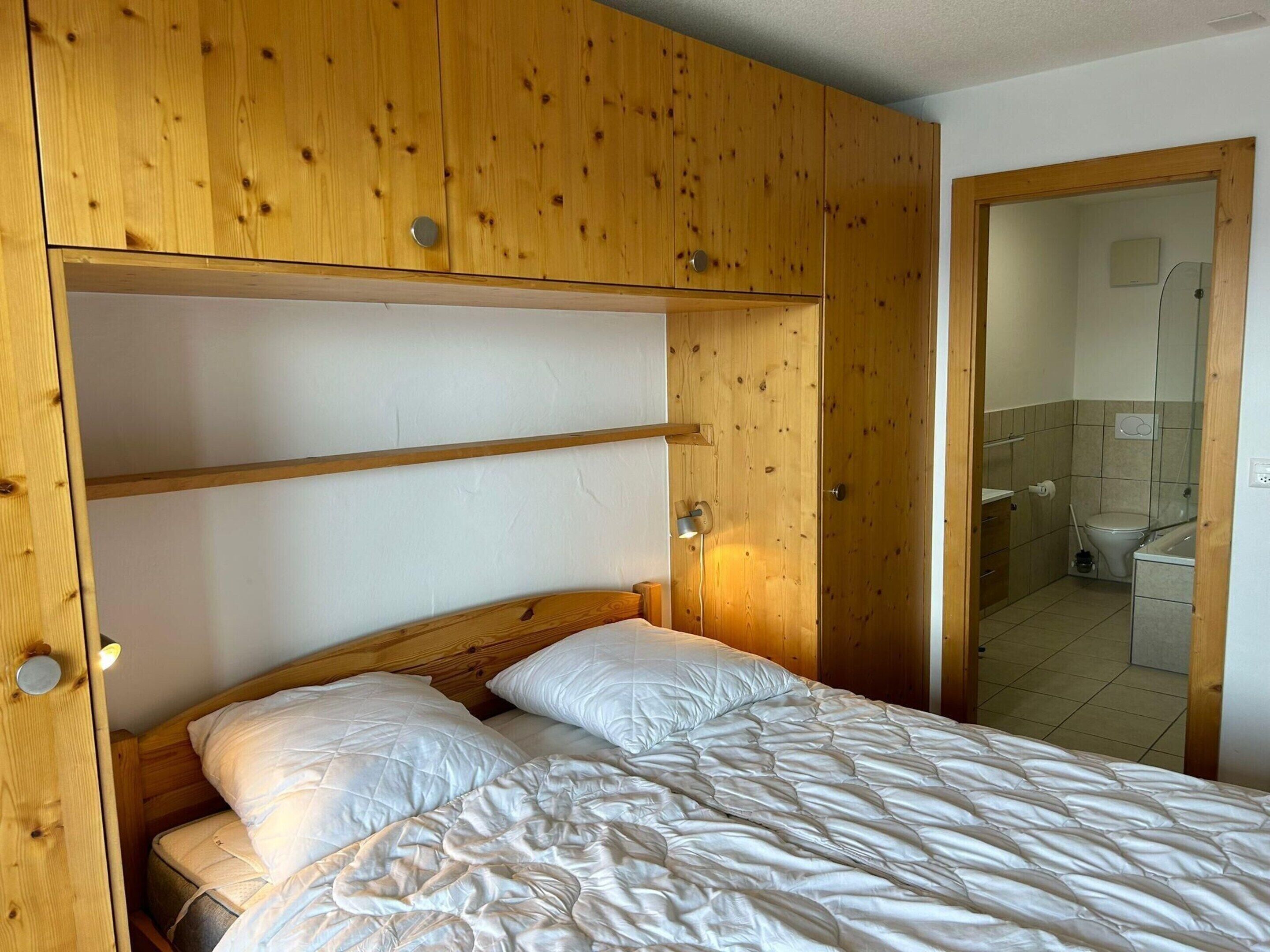 A bedroom with a wooden wardrobe, a bed with white bedding, and a view into a bathroom.
