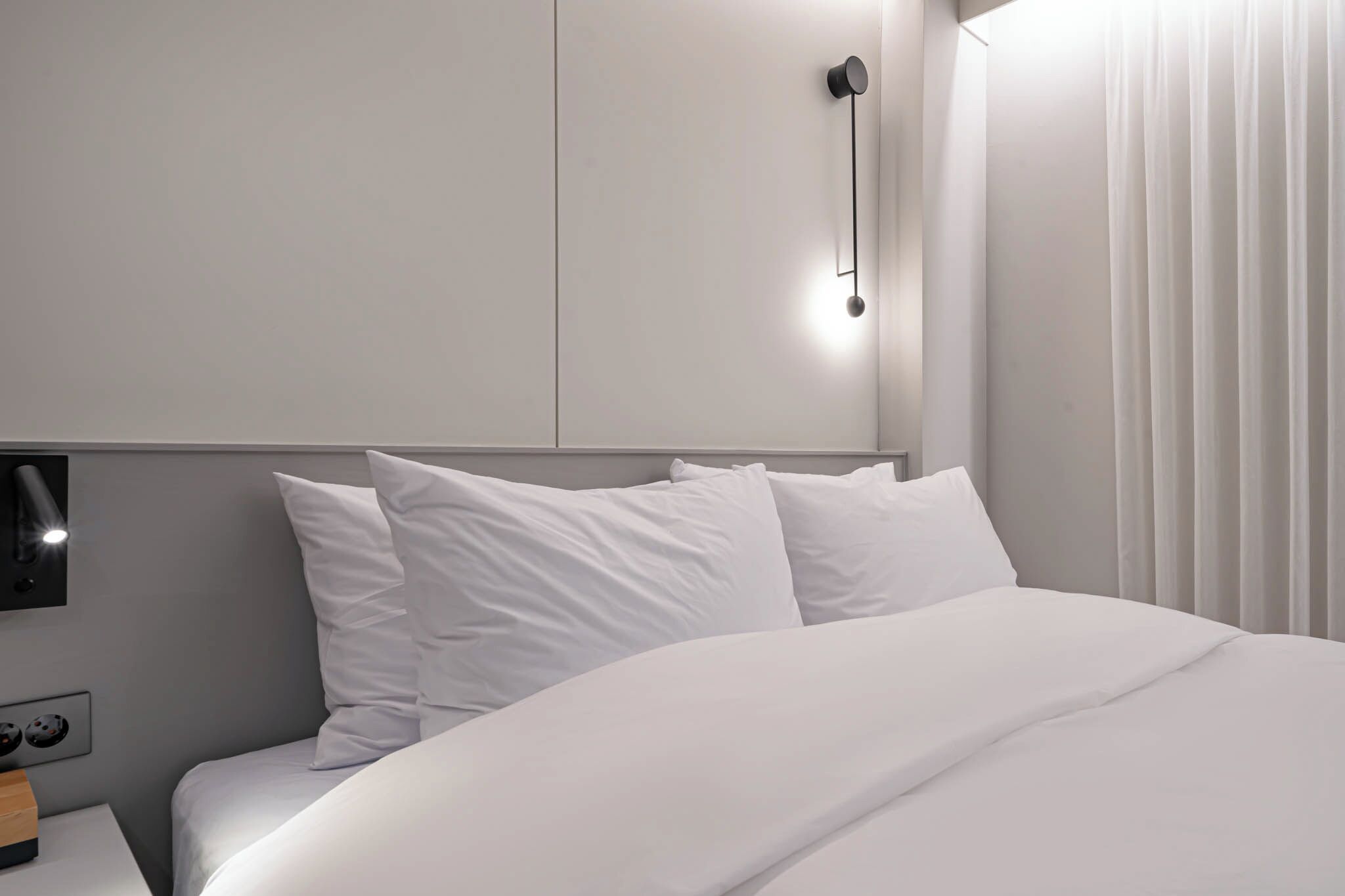 Une chambre d’hôtel moderne avec un lit soigneusement fait, un luminaire fixé au mur et une fenêtre avec des rideaux.