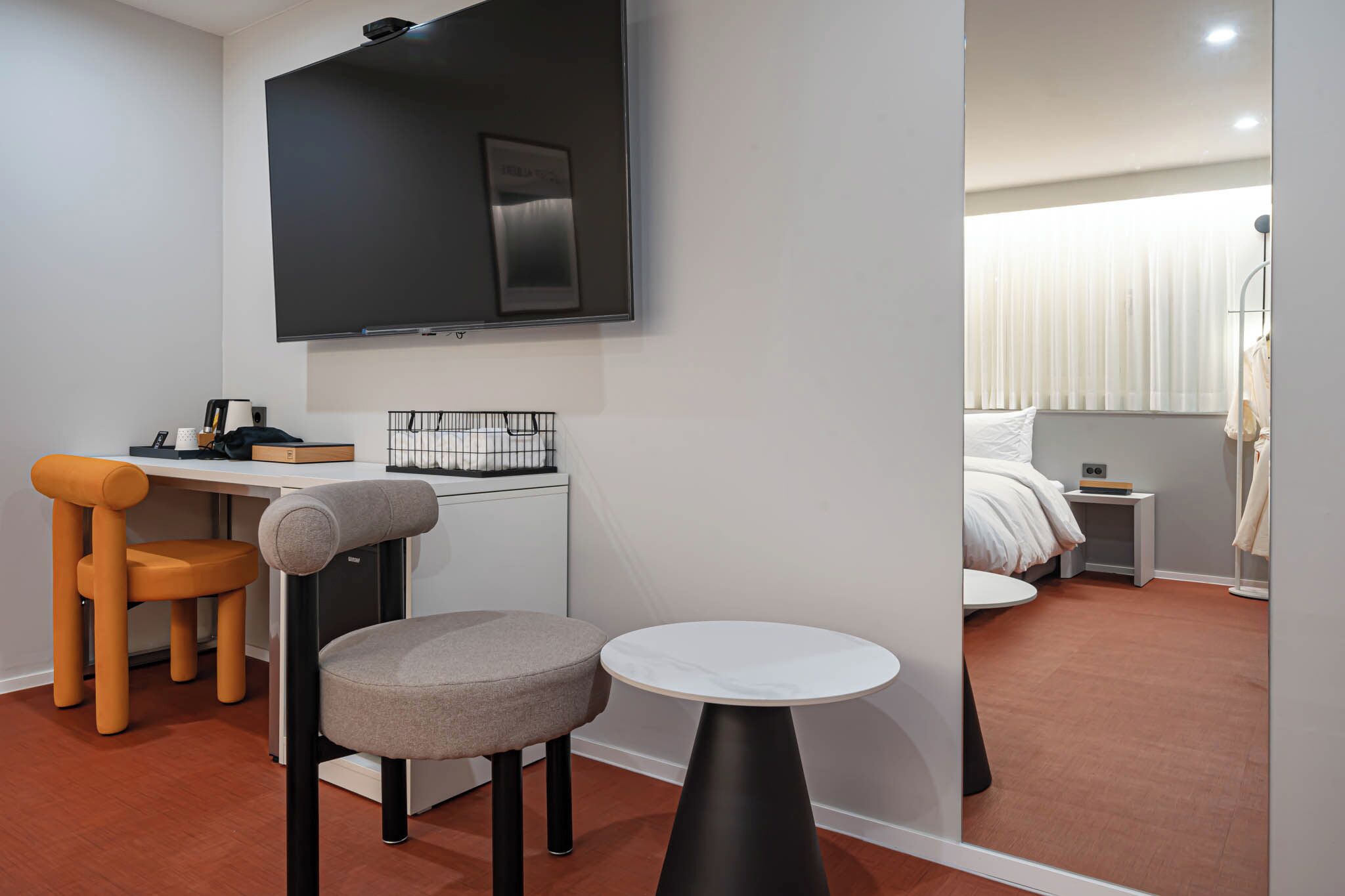 A modern hotel room with a flat-screen TV mounted on the wall, a small round table, two chairs, a bed with white bedding, a nightstand with a radio, and a window with curtains.