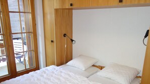 A bedroom with a wooden headboard, two beds with white bedding, and a window with a view of outdoor furniture. - Apartment With Mountain Views (Sion)