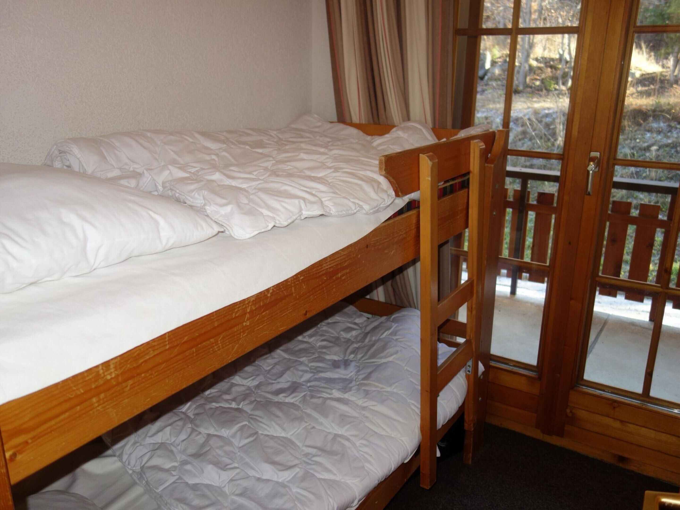 A bunk bed with white bedding in a room with a wooden door and a view of the outdoors.