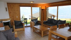 A cozy living room with wooden furniture, a fireplace, and large windows offering a view of the outdoors. - Authentic Apartment With Mountain Views (Veysonnaz)