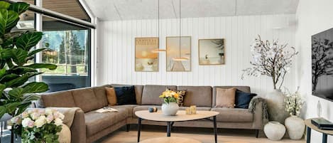 A modern living room with a sectional sofa, round coffee table, and two ottomans. There are large windows, a ceiling with exposed beams, and wall-mounted artwork.
