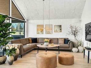 Uma sala de estar moderna com sofá em forma de L, mesa de centro redonda e dois pufes. Há janelas amplas, teto com vigas aparentes e obras de arte fixadas na parede.