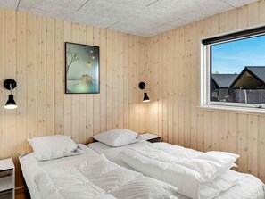 Un dormitorio con paredes de madera, una cama con ropa blanca, dos mesitas de noche, una ventana con vista a casas y un cuadro en la pared.
