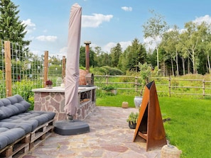 Een terras buiten met een stenen open haard, een overdekte barbecue en een zithoek met kussens.