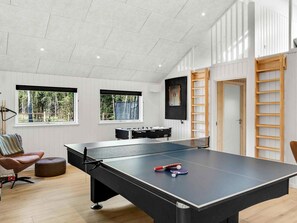 Mesa de ping pong con red, paleta de tenis de mesa y pelota en una habitación con piso de madera, una silla acolchada y ventanas con vista a árboles.