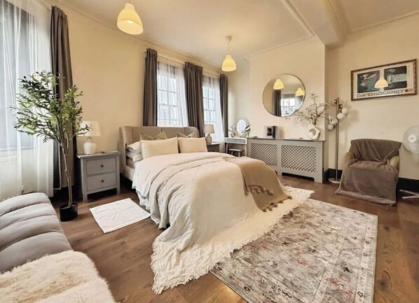 Une chambre à coucher comprenant un lit, des tables de chevet, un bureau, une chaise, un miroir, une plante en pot et une grande fenêtre avec des rideaux.