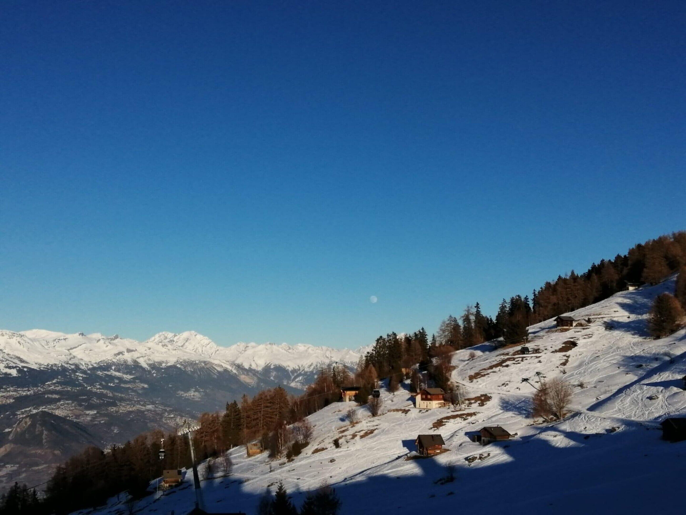 A snow-covered mountain landscape with a clear sky, a few buildings, and a visible moon.