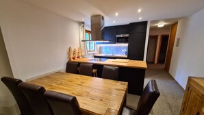A compact kitchen with a central island, dark cabinetry, and a dining area with a wooden table and chairs. - Radiant Apartment Facing Hills (Veysonnaz)