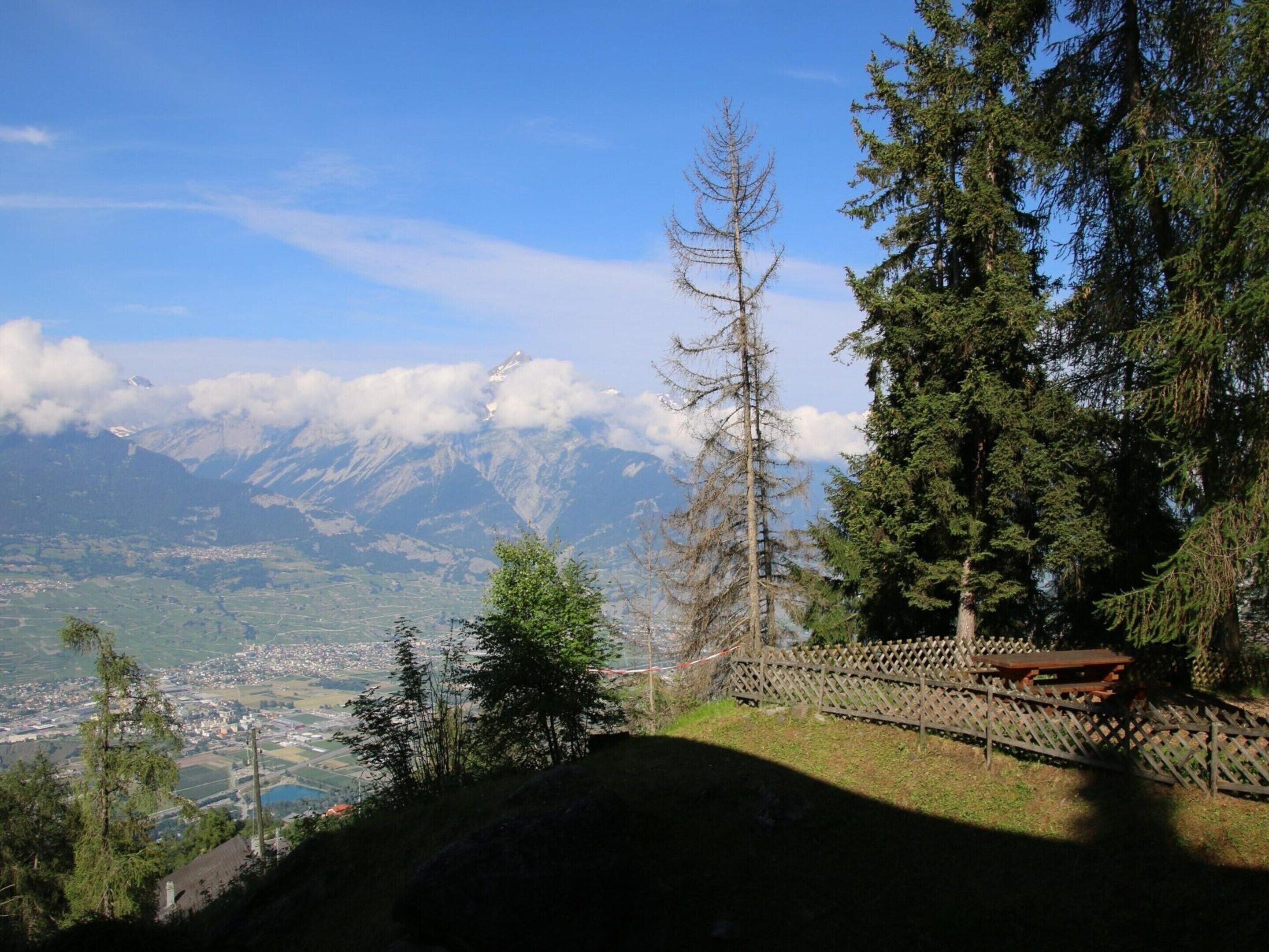 Un paysage montagneux offrant une vue dégagée sur une ville située en contrebas, avec une clôture en bois et une table de pique-nique.