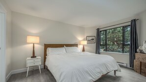 A neatly arranged bedroom with a large bed, two bedside tables with lamps, a window with curtains, and a framed picture on the wall. - Live the North Shore dream in this mountain-view penthouse! (North Vancouver)
