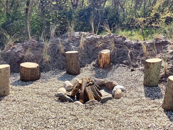 A natural outdoor setting with a fire pit surrounded by rocks and logs, with trees and grass in the background.