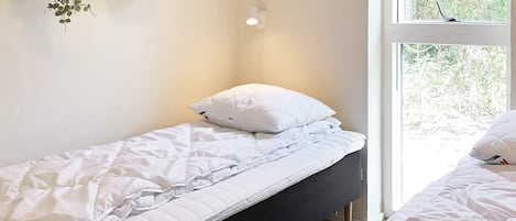A single bed with white bedding, a bedside table, and a window with a view of greenery.