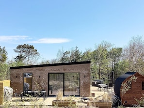 Ein modernes, eingeschossiges Gebäude mit großen Glasfenstern, einer Terrasse mit Gartenmöbeln und einem Holzboden.