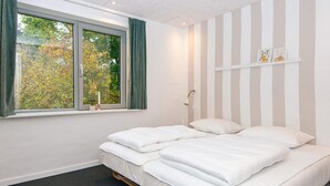 Een slaapkamer met een groot raam, een bed met witte lakens, een nachtkastje en een wandplank met boeken.