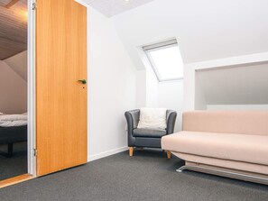 Et værelse med dør i træ, en grå lænestol, en beige sofa, et lille vindue og et skråt loft.