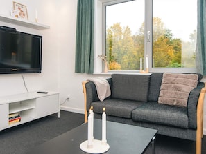 A modern living room with a flat-screen TV, a grey sofa, a coffee table with candles, and a window with a view of autumn trees. - 6 Person Holiday Home in Ansager (Ansager)